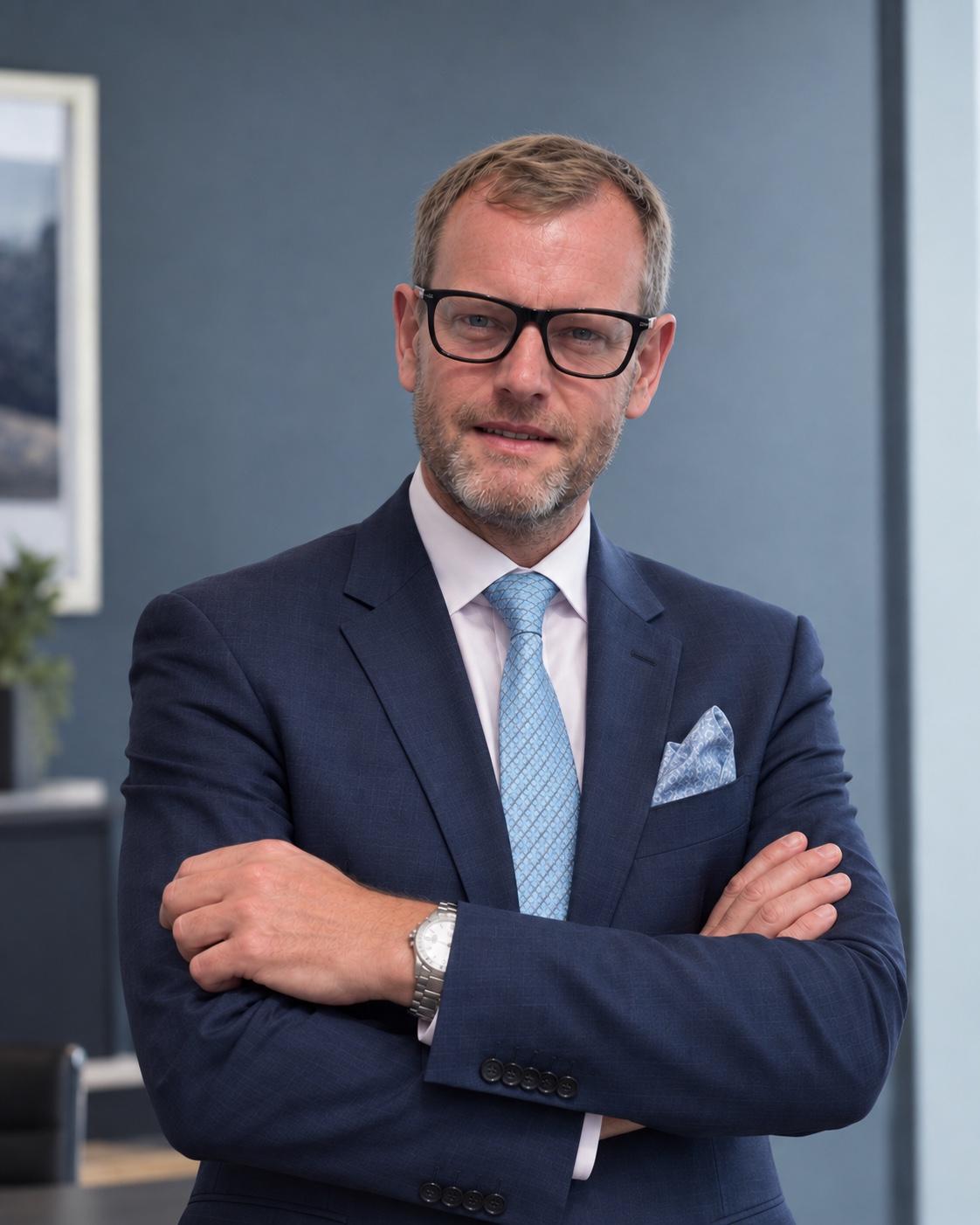Will McKintosh, Founder & Principal of Saanen Private Office, in a navy suit and light blue tie, arms folded, against a dark blue-grey wall.